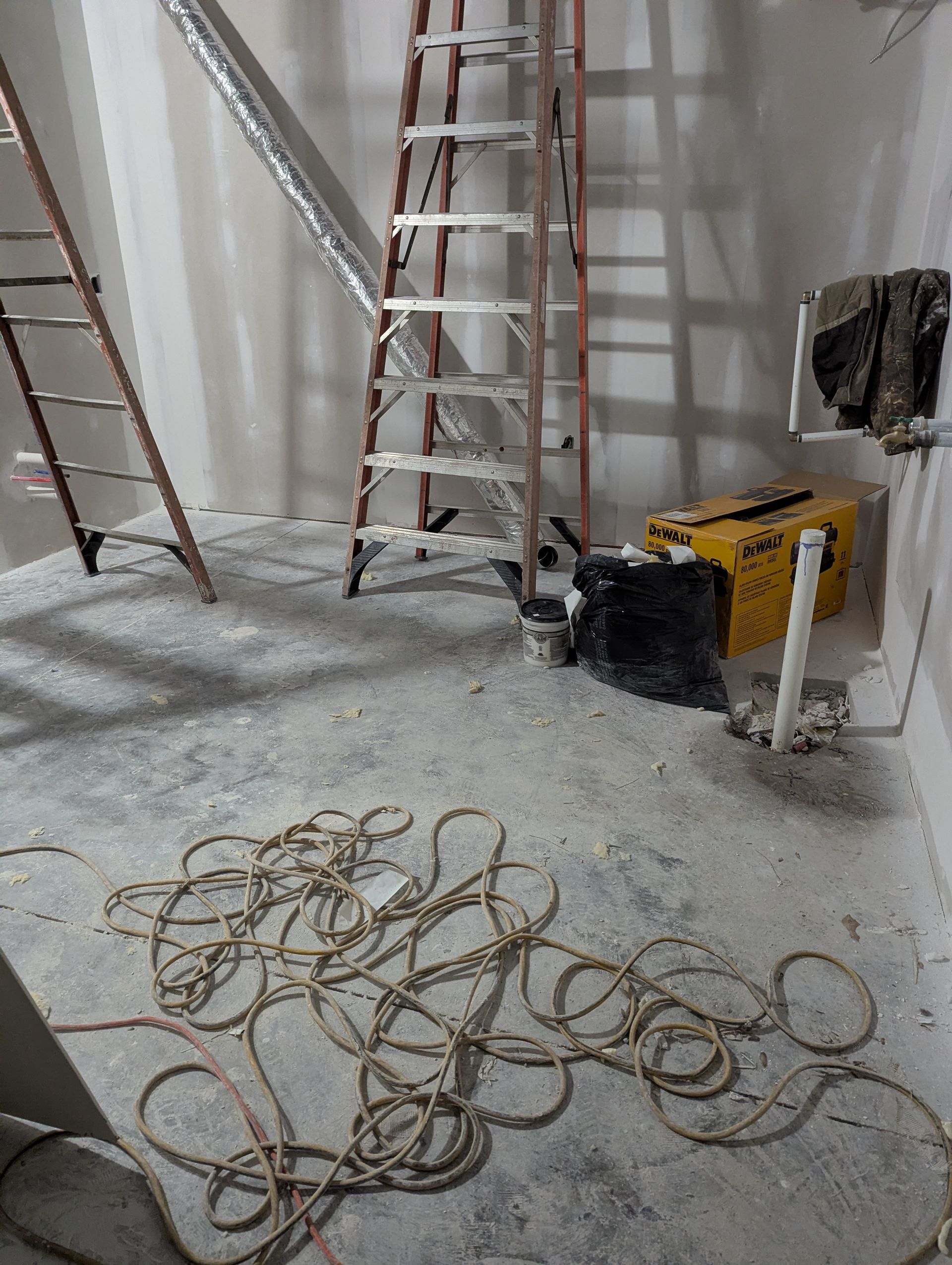 A construction site room with two ladders, a yellow box, and a coiled electrical cord on a dusty concrete floor.