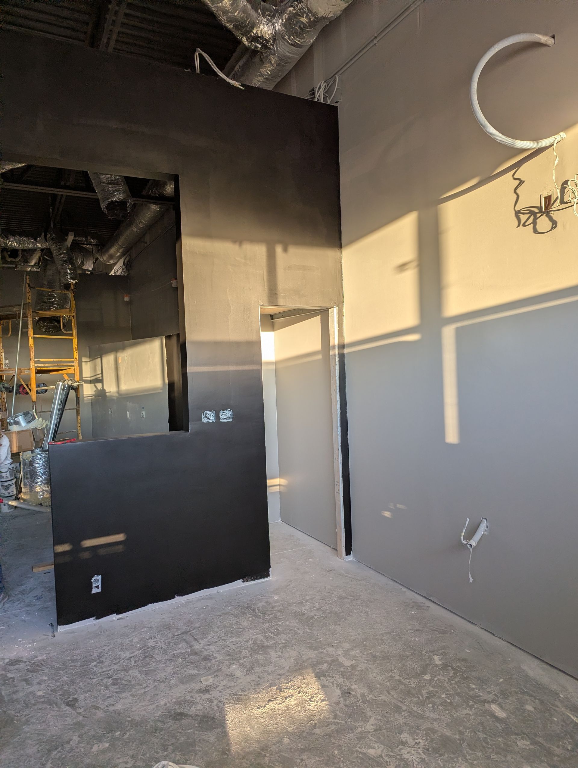 Construction site interior with gray and black walls, an open doorway, exposed ceiling pipes, and natural light.