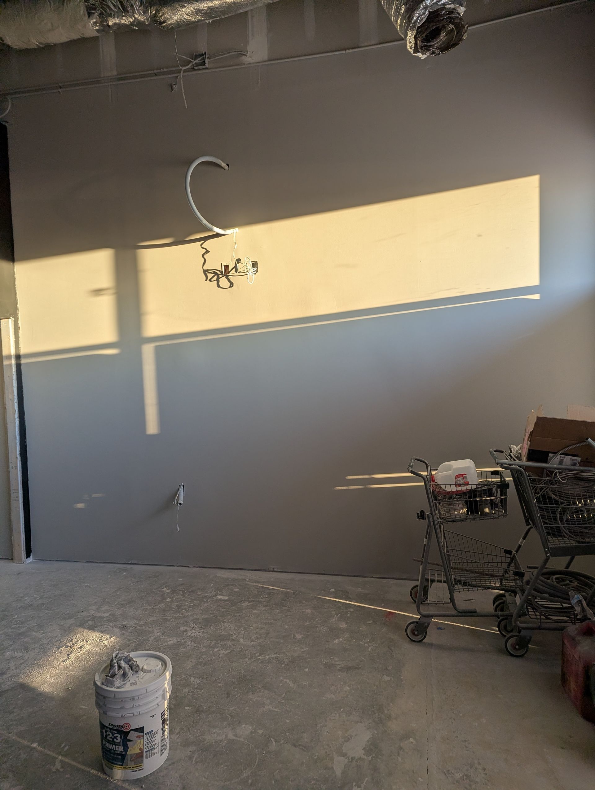 Sunlight streams through a window onto a grey wall in an unfinished room with a construction bucket and shopping cart.