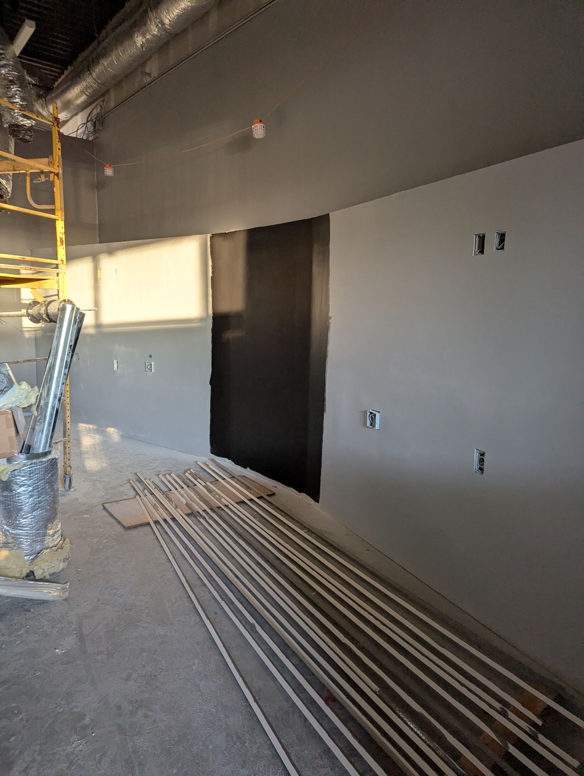 A construction site showing a gray wall with a black patch, exposed ductwork, a yellow scaffold, and metal trim on the floor.