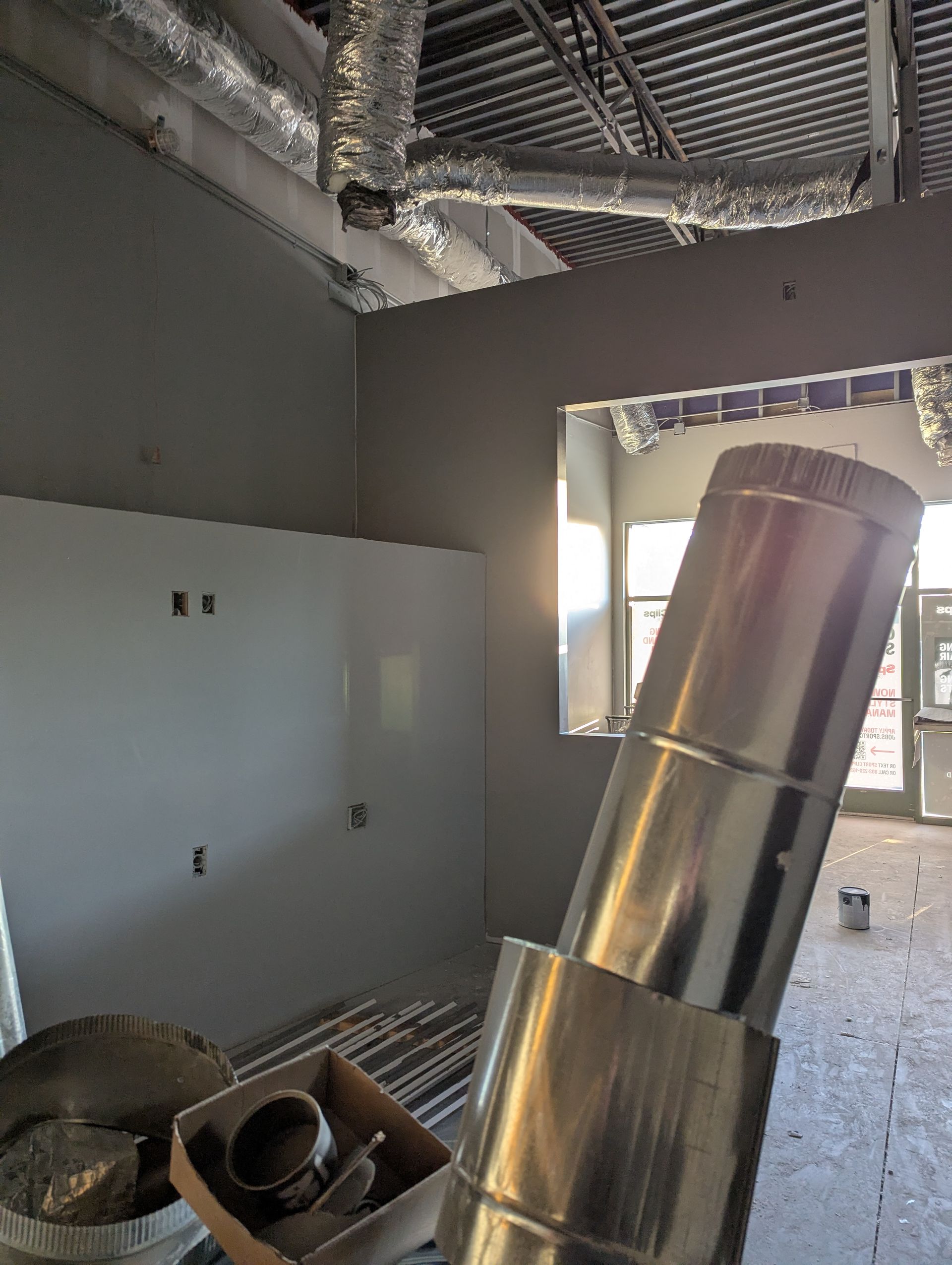 Interior view of a construction site showing exposed ceiling ducts, grey walls, a doorway, and a metal duct pipe foreground.