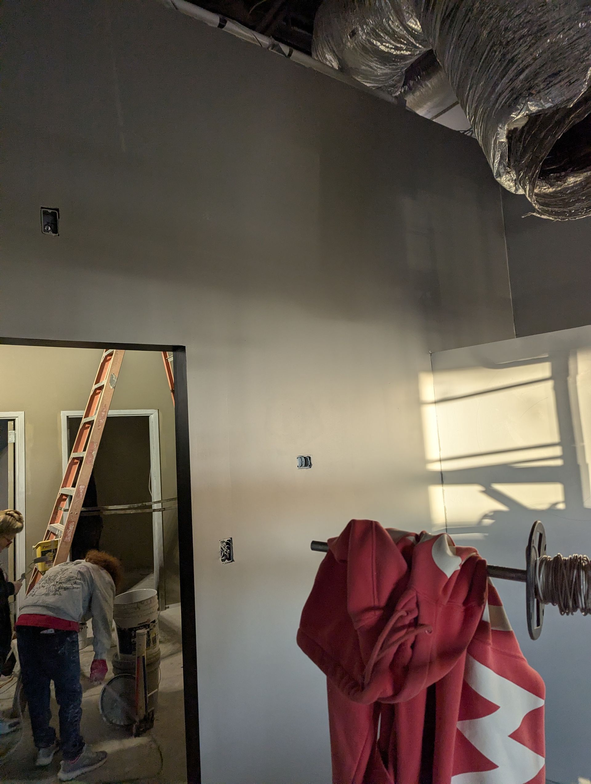 A construction worker climbs a ladder in a room with unfinished drywall, exposed ductwork, and hanging red fabric.