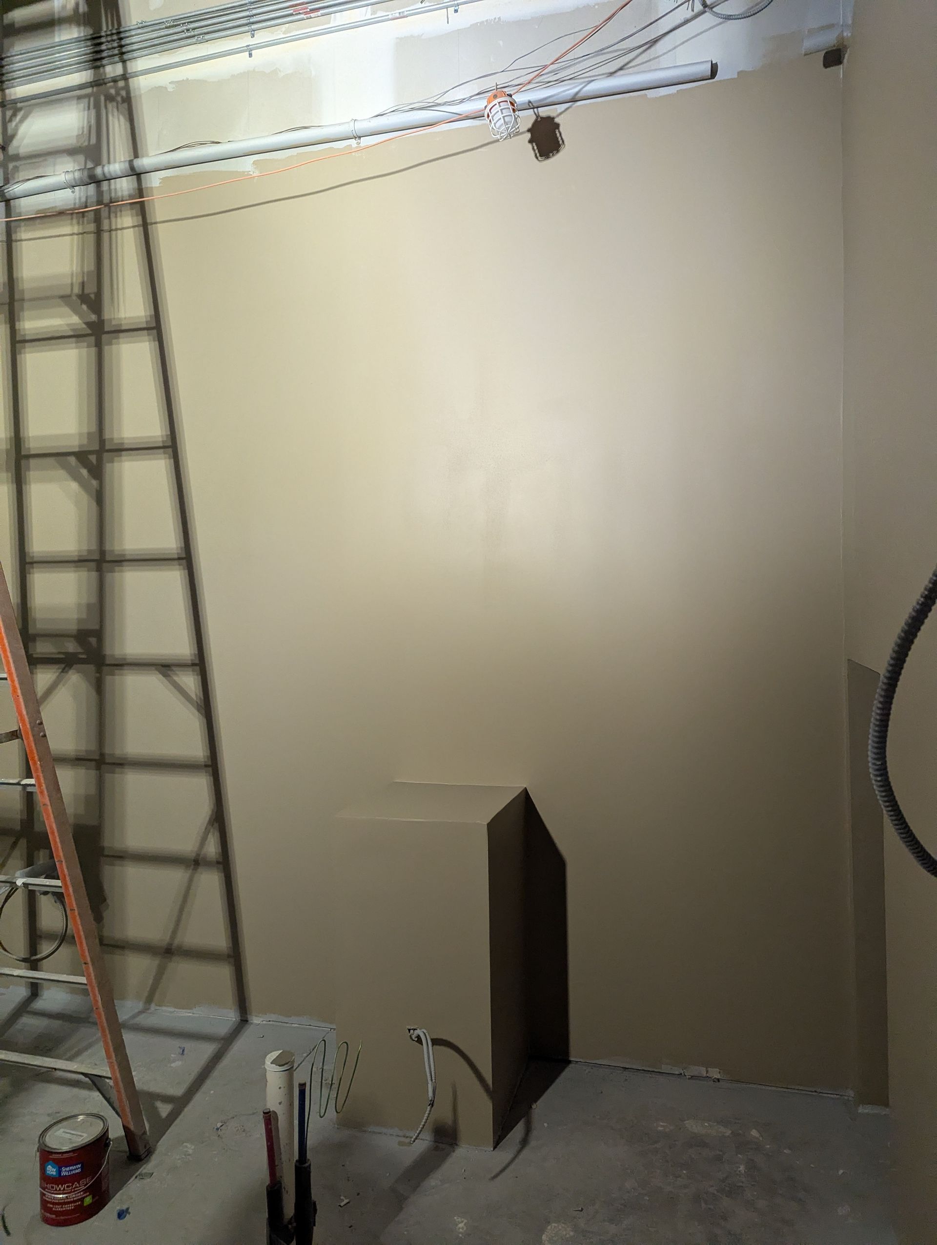 A ladder stands against a freshly painted tan wall in an unfinished room with visible wires near the ceiling.