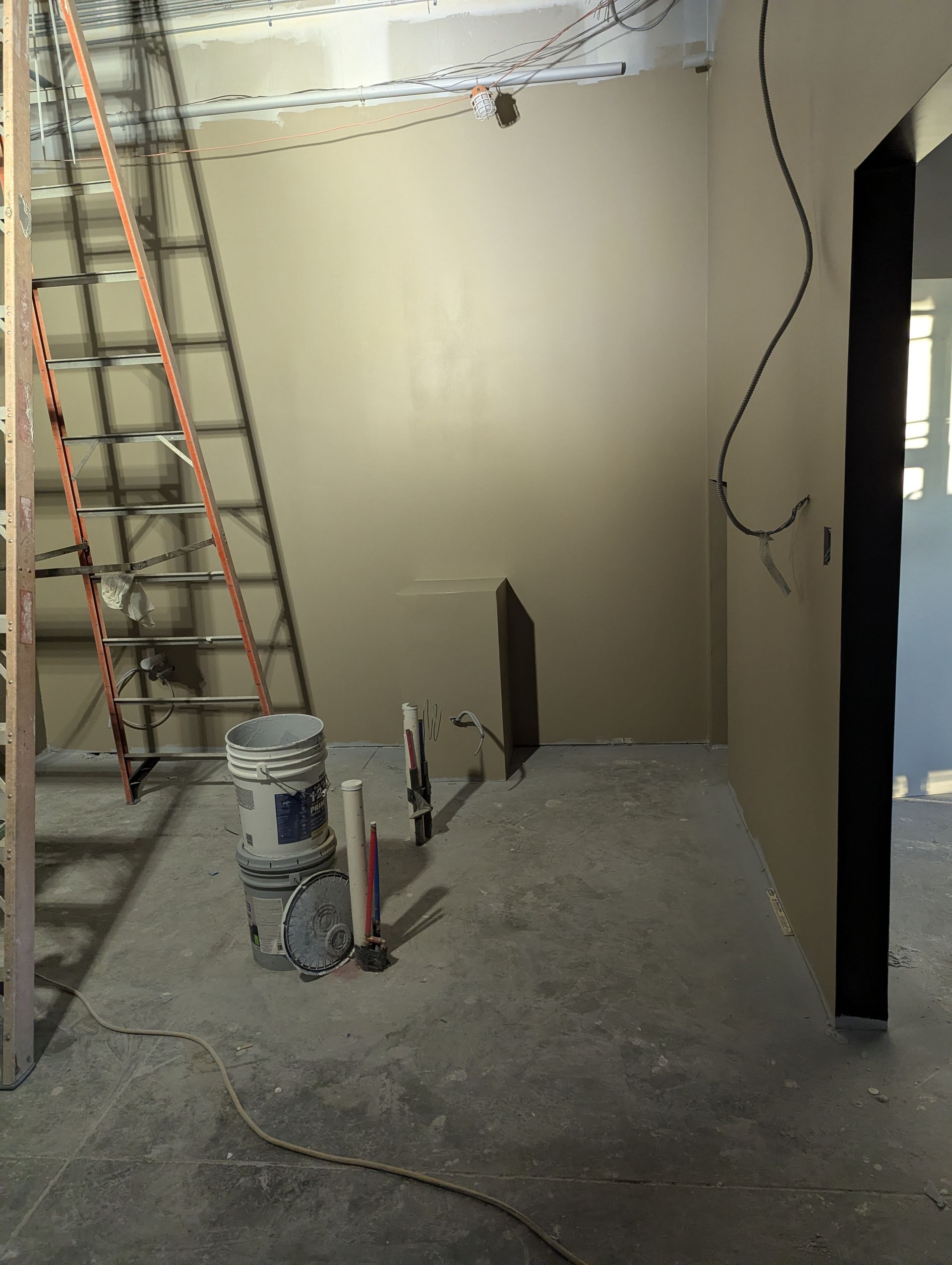 An unfinished room with beige walls, a ladder on the left, exposed wires, and a paint bucket on the concrete floor.