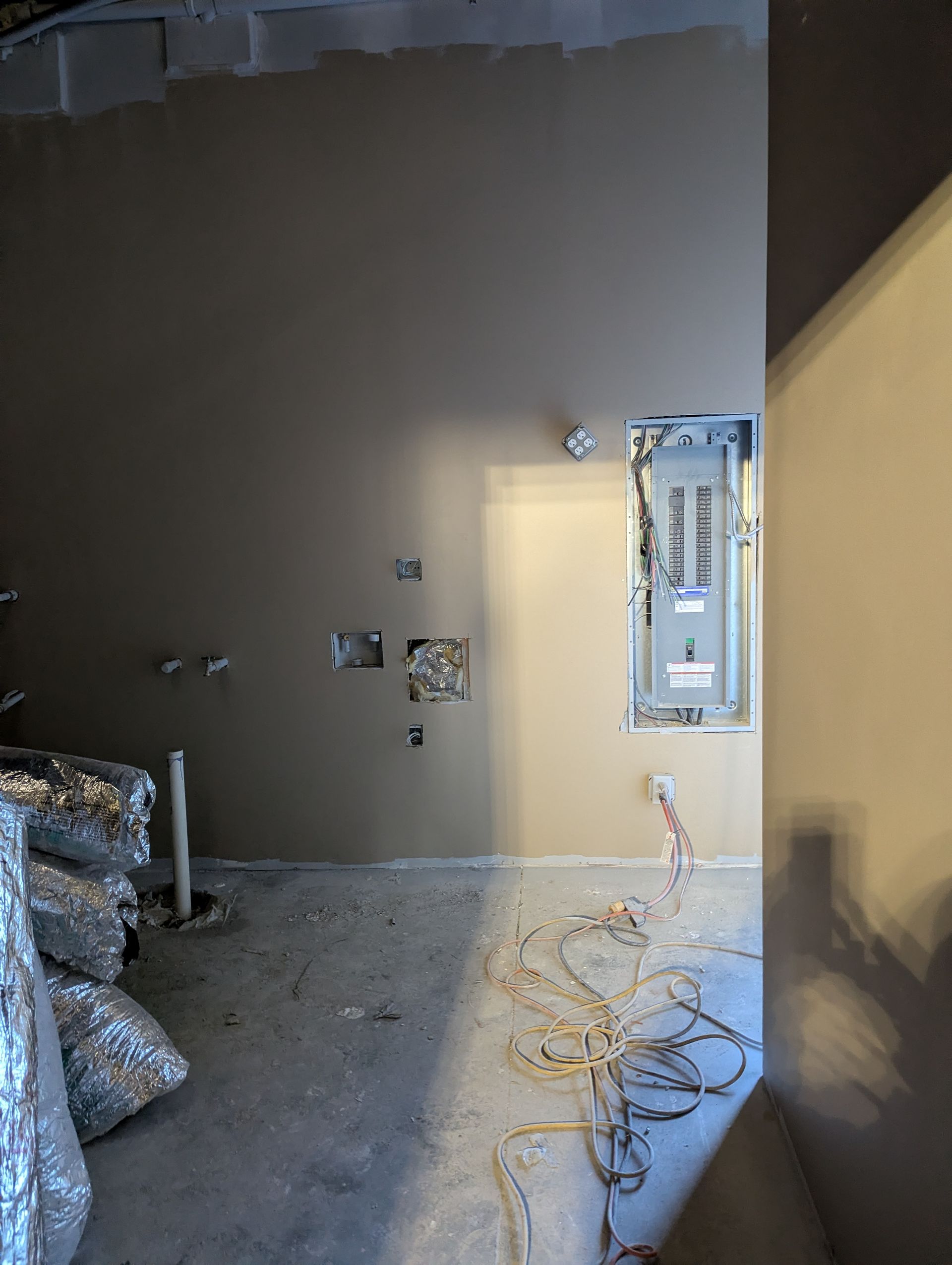 An unfinished room interior with an electrical panel, exposed wiring, and wall outlets on a beige wall.