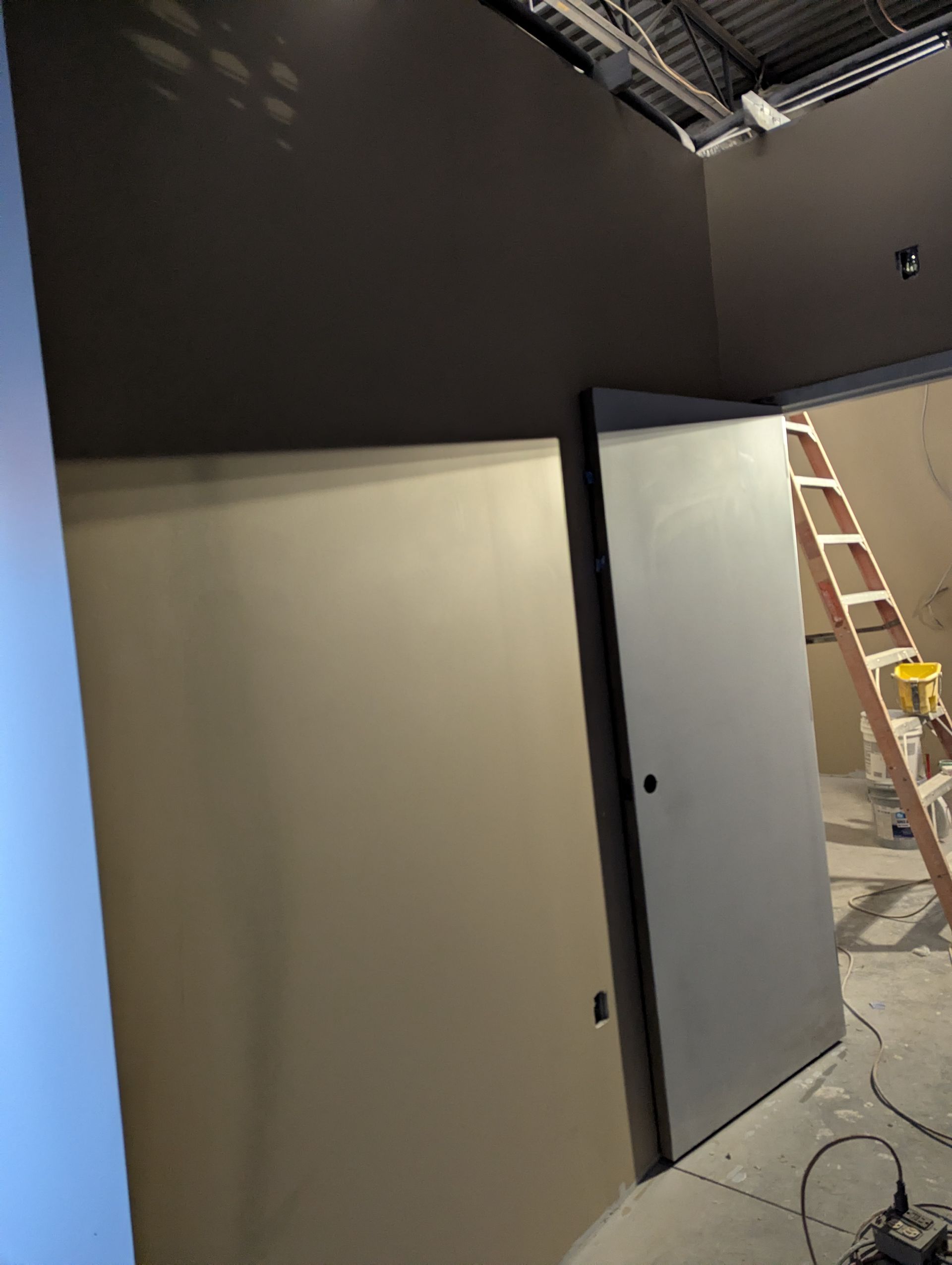 Interior construction site showing a dark brown wall above a light tan wall, a gray door, and a wooden ladder.
