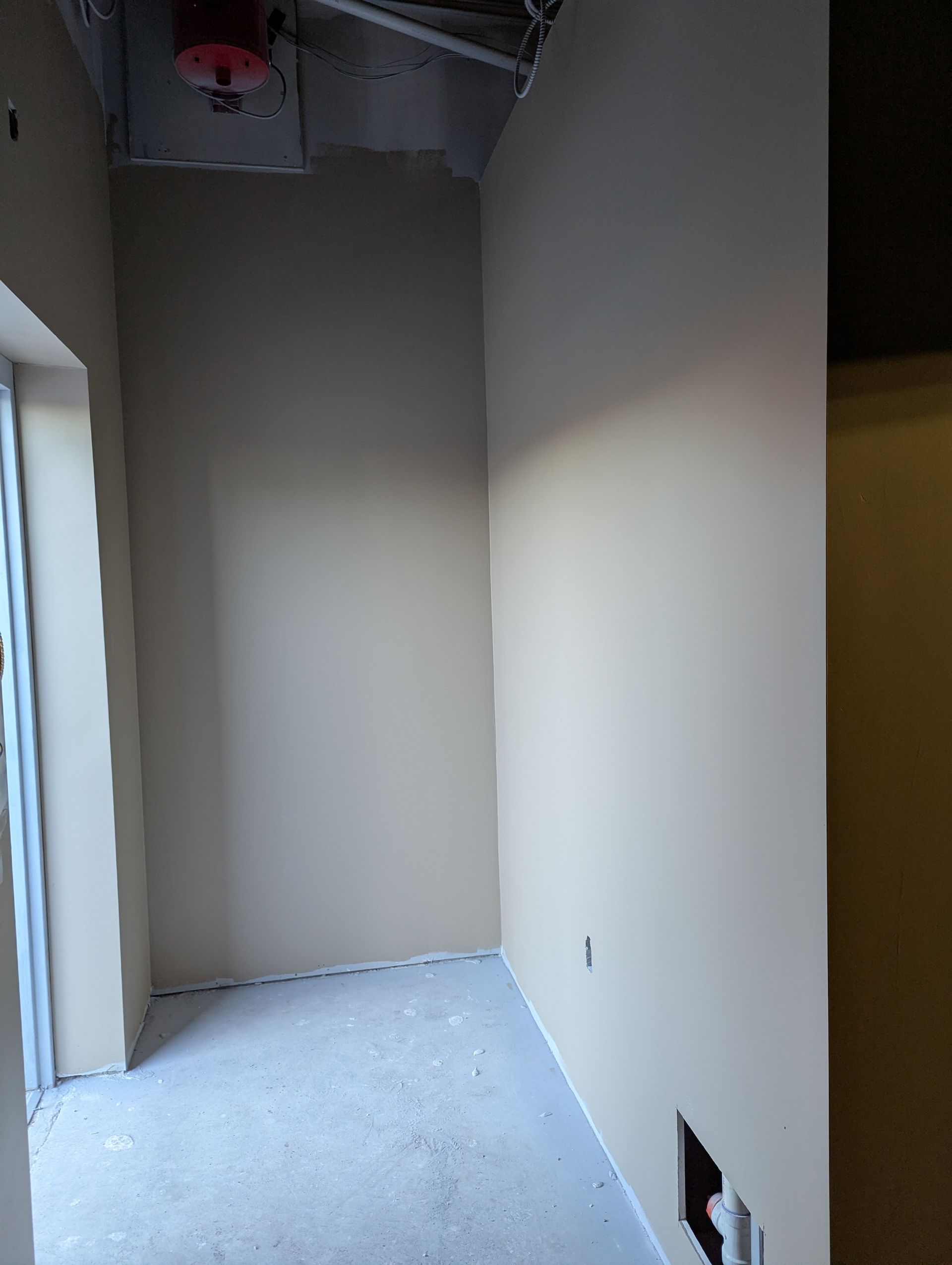 An unfinished, narrow room with beige walls, an exposed ceiling structure with a red object, and a concrete floor.