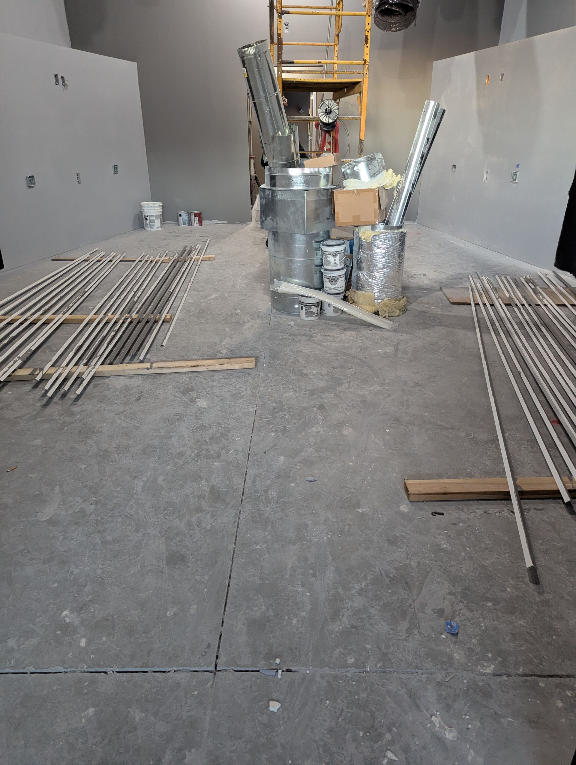 Construction site with drywall panels, metallic HVAC ductwork, and bundles of metal rods on a concrete floor.