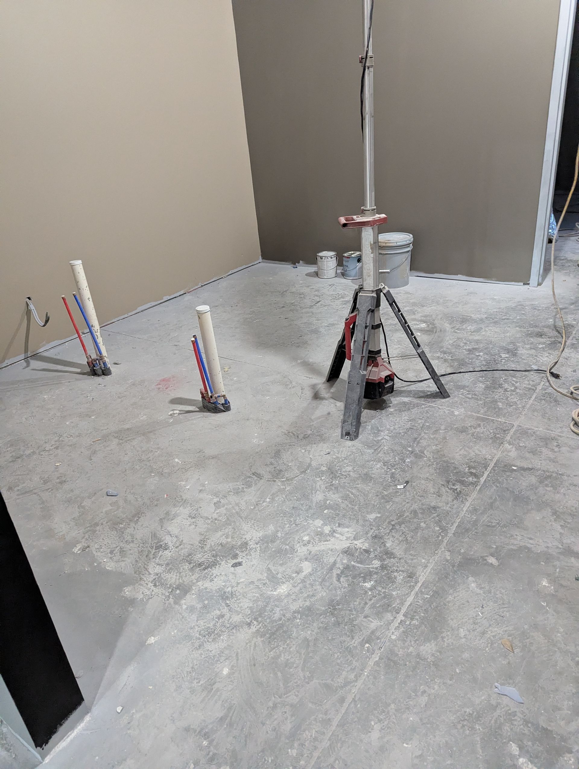 Interior construction site with plumbing pipes protruding from a concrete floor and a tripod-mounted tool stand.