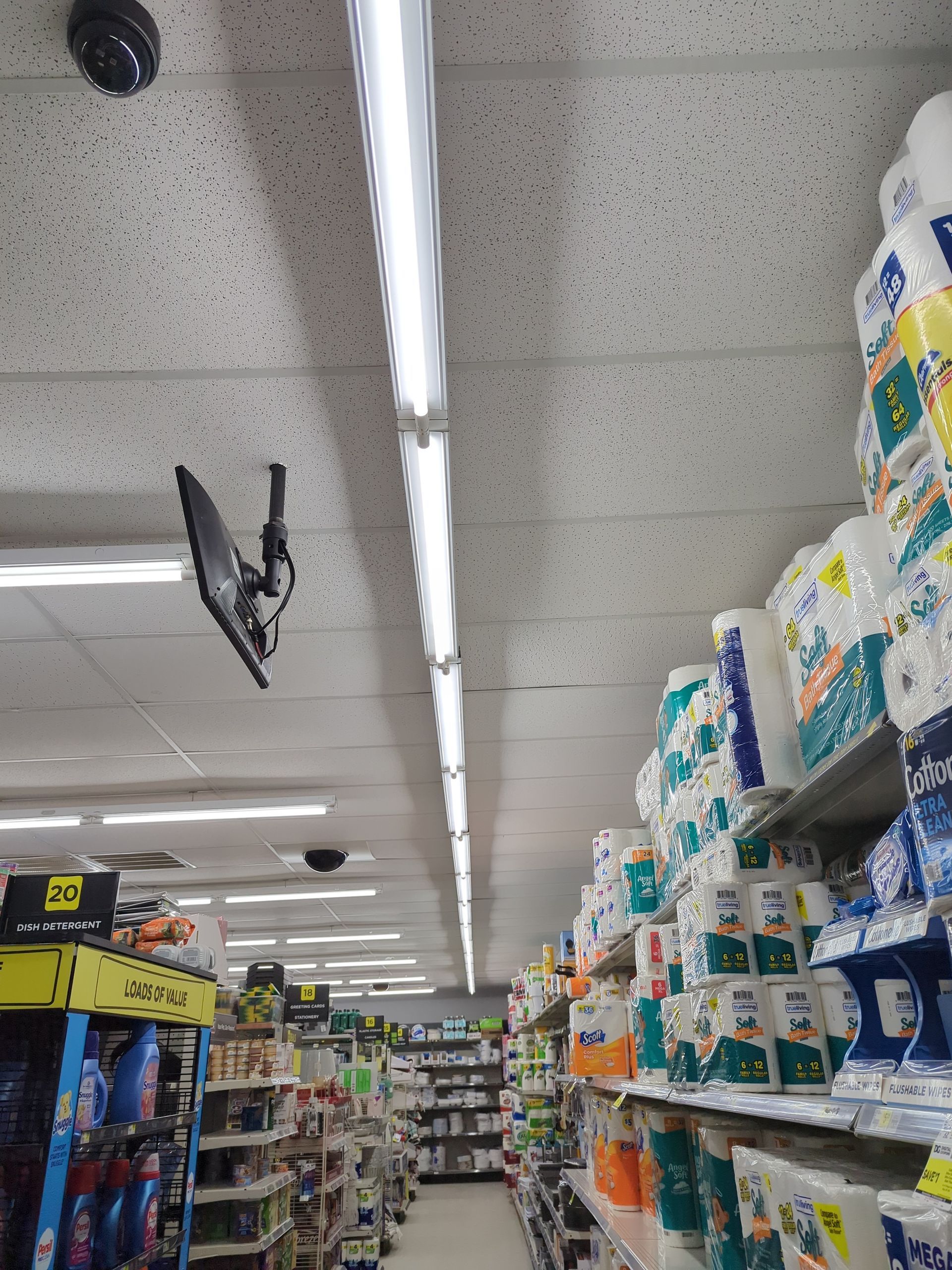 A low-angle view of a retail aisle lined with shelves of paper products under bright fluorescent ceiling lights.