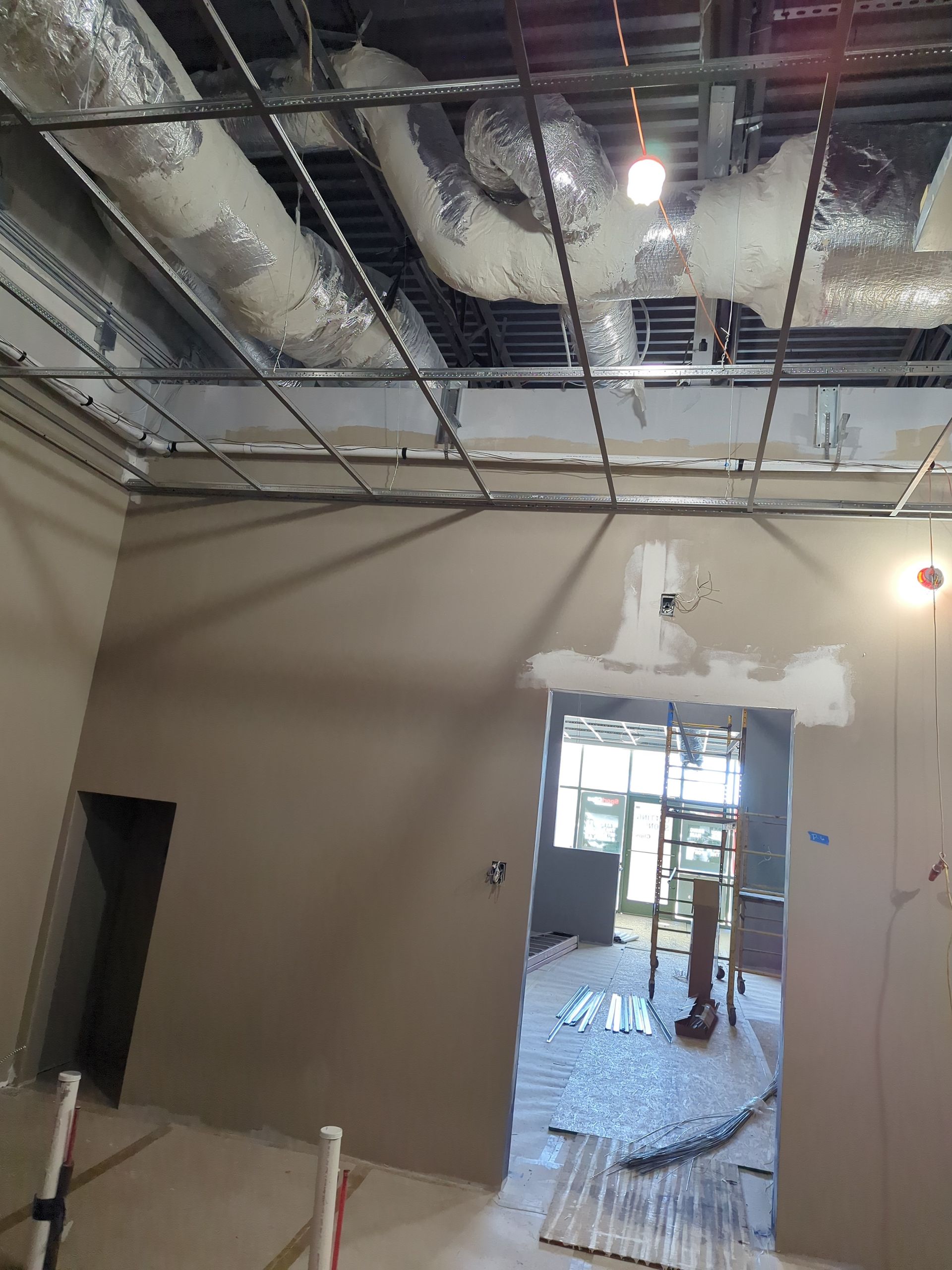 An interior construction site with exposed ceiling infrastructure, unfinished beige walls, and a doorway leading to light.