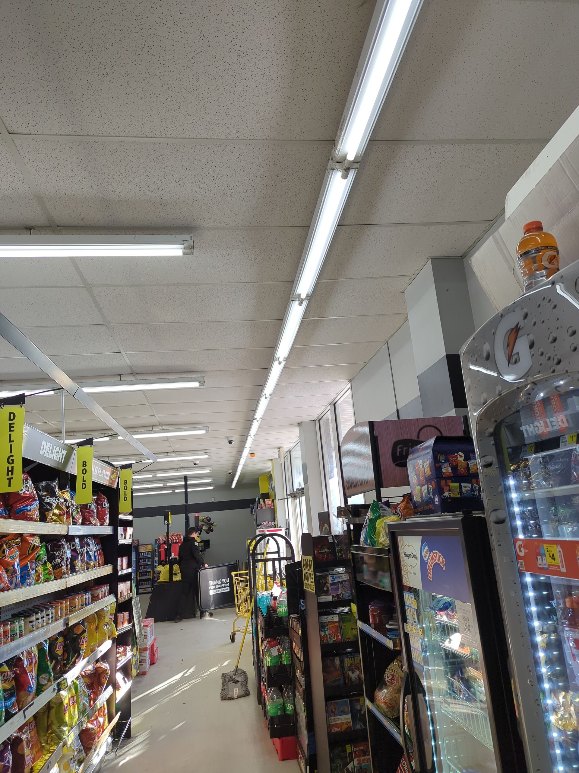 An aisle inside a convenience store with shelves stocked with snacks and drinks, illuminated by overhead lighting.