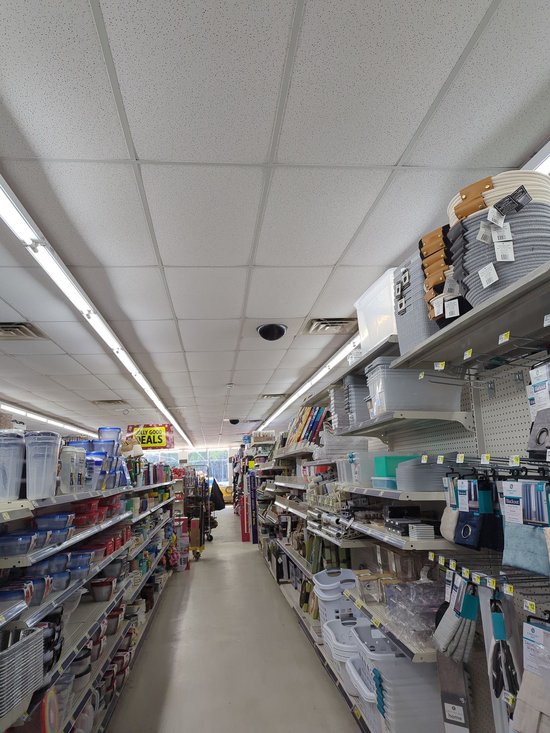 A view down an aisle in a store, with shelves lined with various household goods, clear floors, and bright overhead lights.