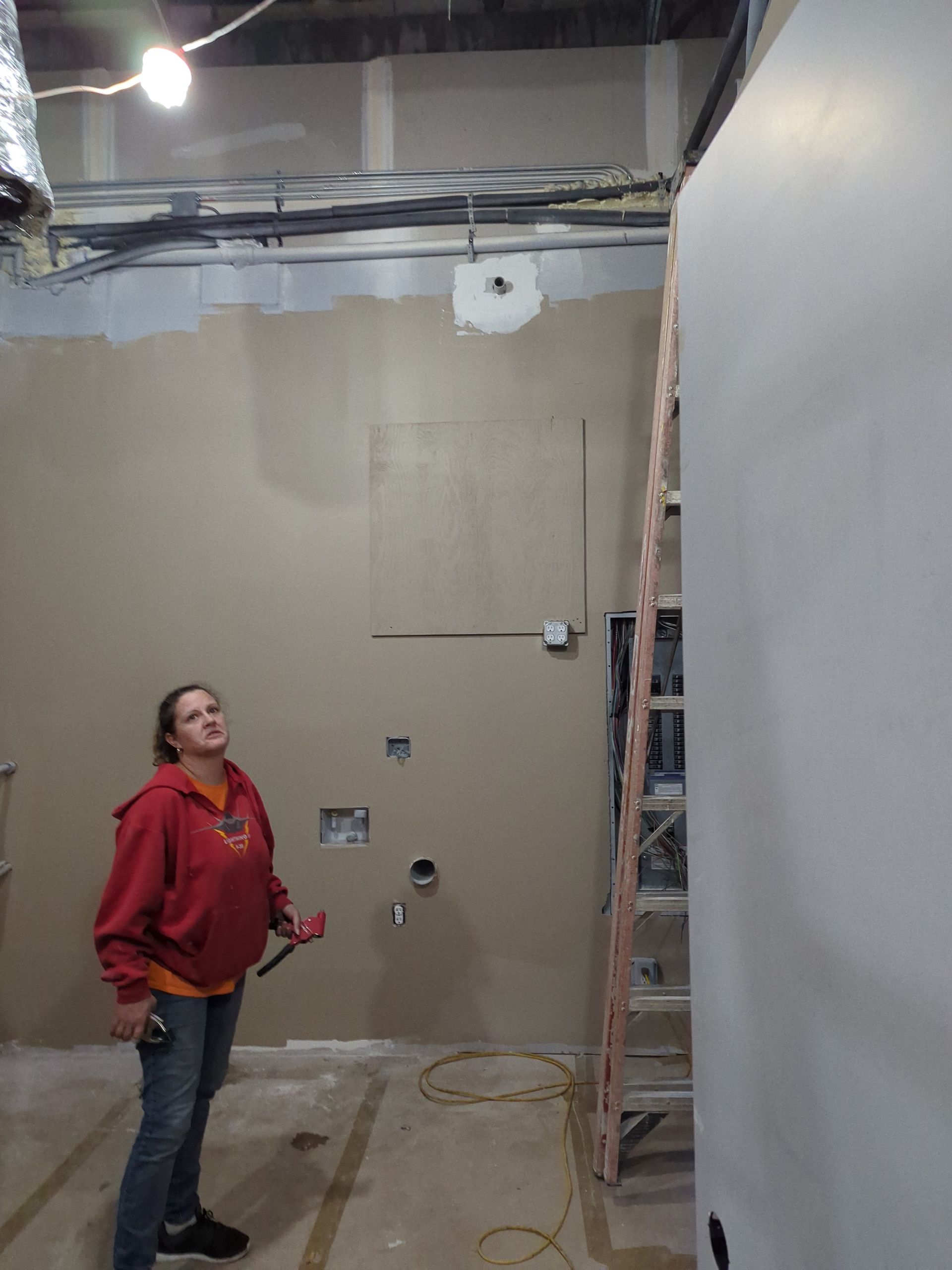 A person in a red sweatshirt holding a tool stands in an unfinished room with drywall and a ladder.