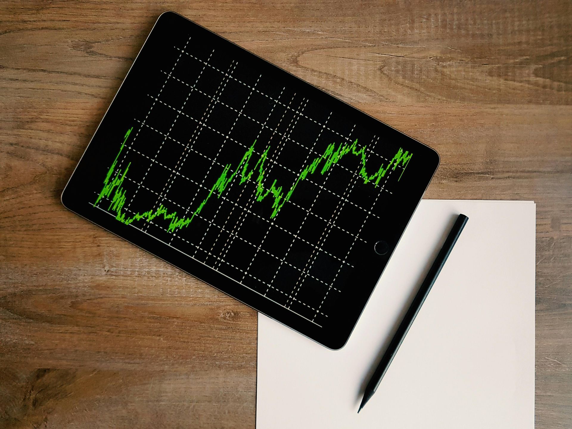 Tablet displaying a green stock chart, with blank paper and pencil on a wood table.