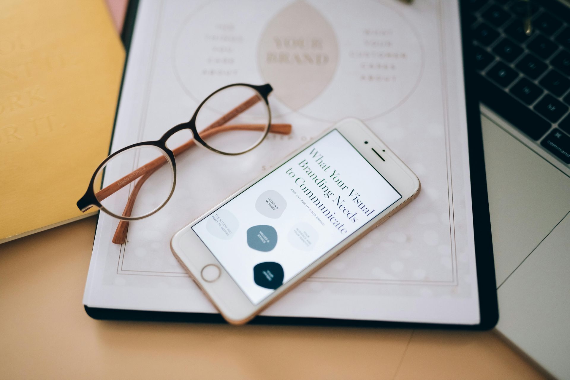 White smartphone, glasses, notebook, and laptop on a desk.