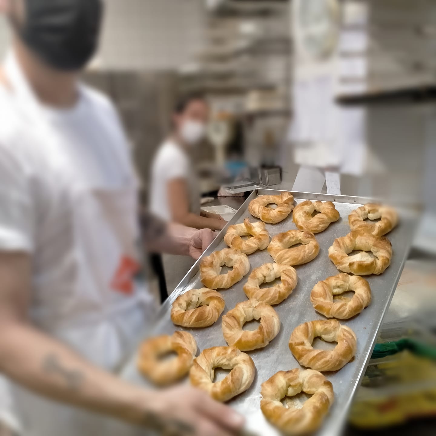 Fornaio che regge un vassoio di dolci appena sfornati. Un altro fornaio sullo sfondo. Ambiente cucina.