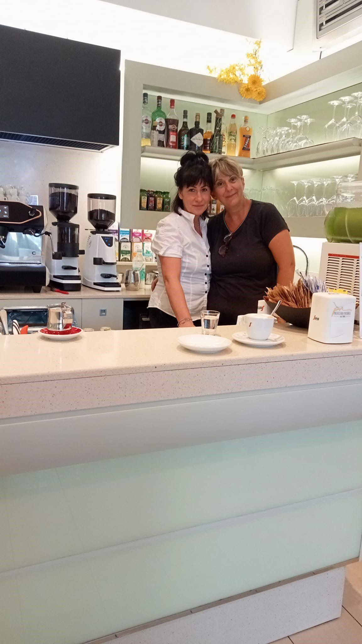 Due donne dietro un bancone di un bar. Una indossa una camicia bianca, l'altra una camicia nera.