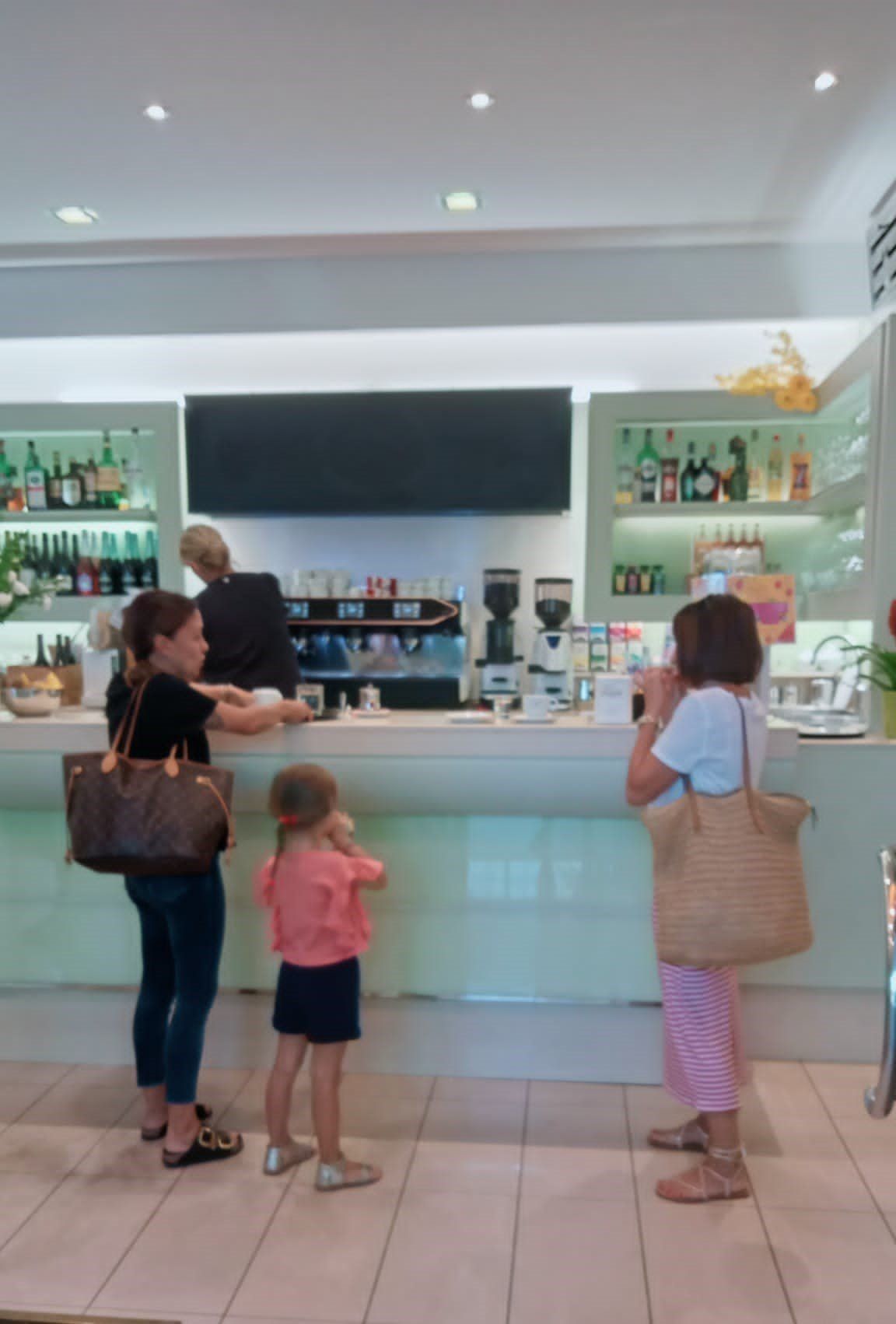 Persone al bancone di un bar. Due donne, un bambino che ordina e una donna che guarda il telefono. Interno luminoso e ben illuminato.