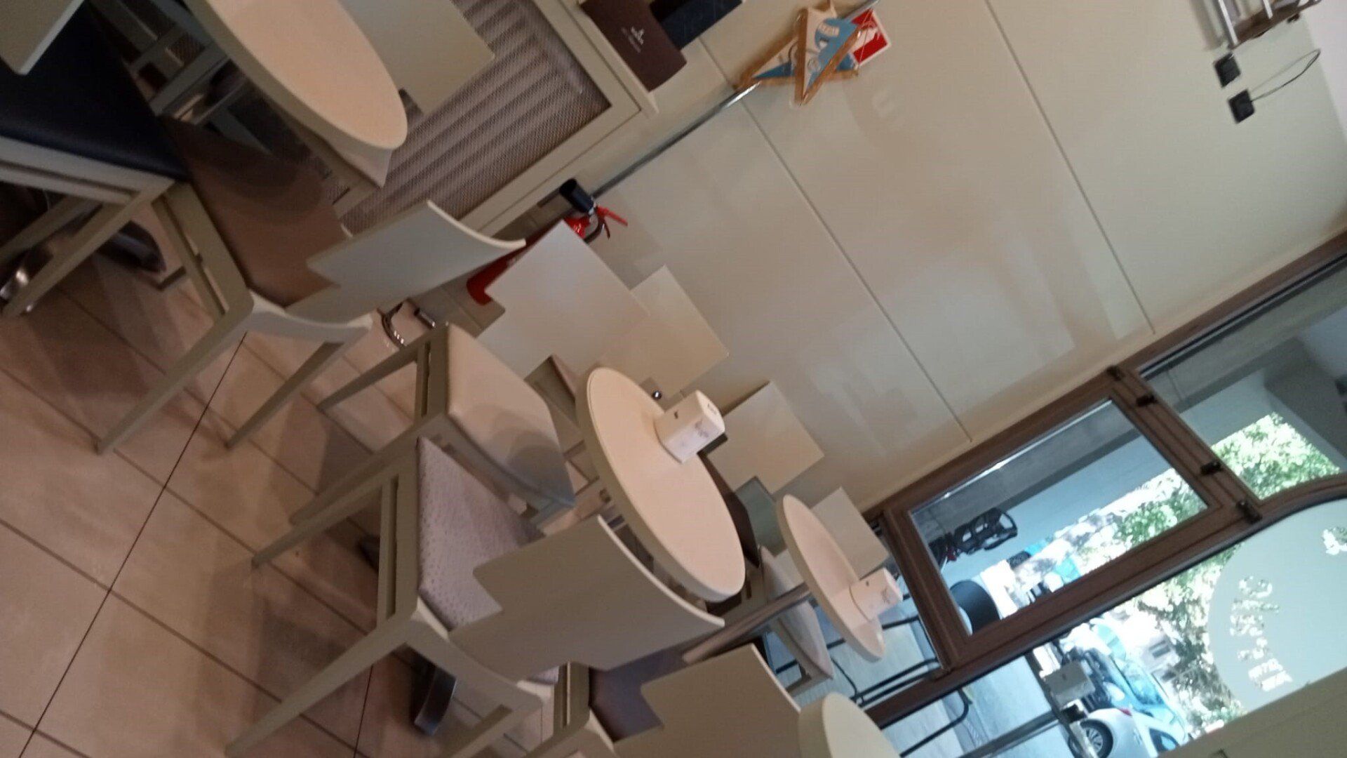 Interno di un bar con tavoli e sedie rotondi bianchi. Una porta a vetri e finestre offrono una vista sull'esterno.