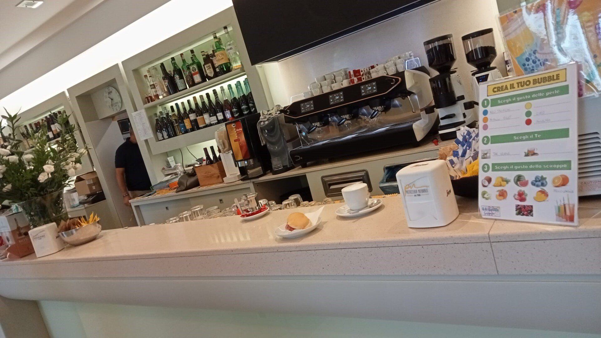Interno di un bar con bancone, macchina per l'espresso e bottiglie; dietro il bancone è visibile una persona.