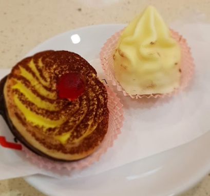 Due pasticcini su un piatto bianco: uno circolare con spirali gialle e cioccolato e una ciliegia in cima, e un cono glassato bianco.