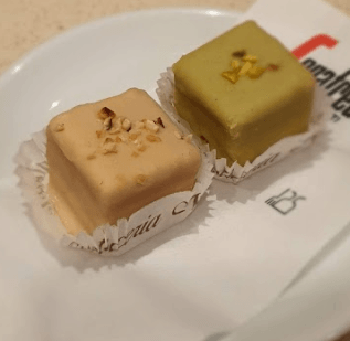 Due piccoli pasticcini quadrati: uno color nocciola e uno verde pistacchio, con sopra delle noci, su un piatto bianco.