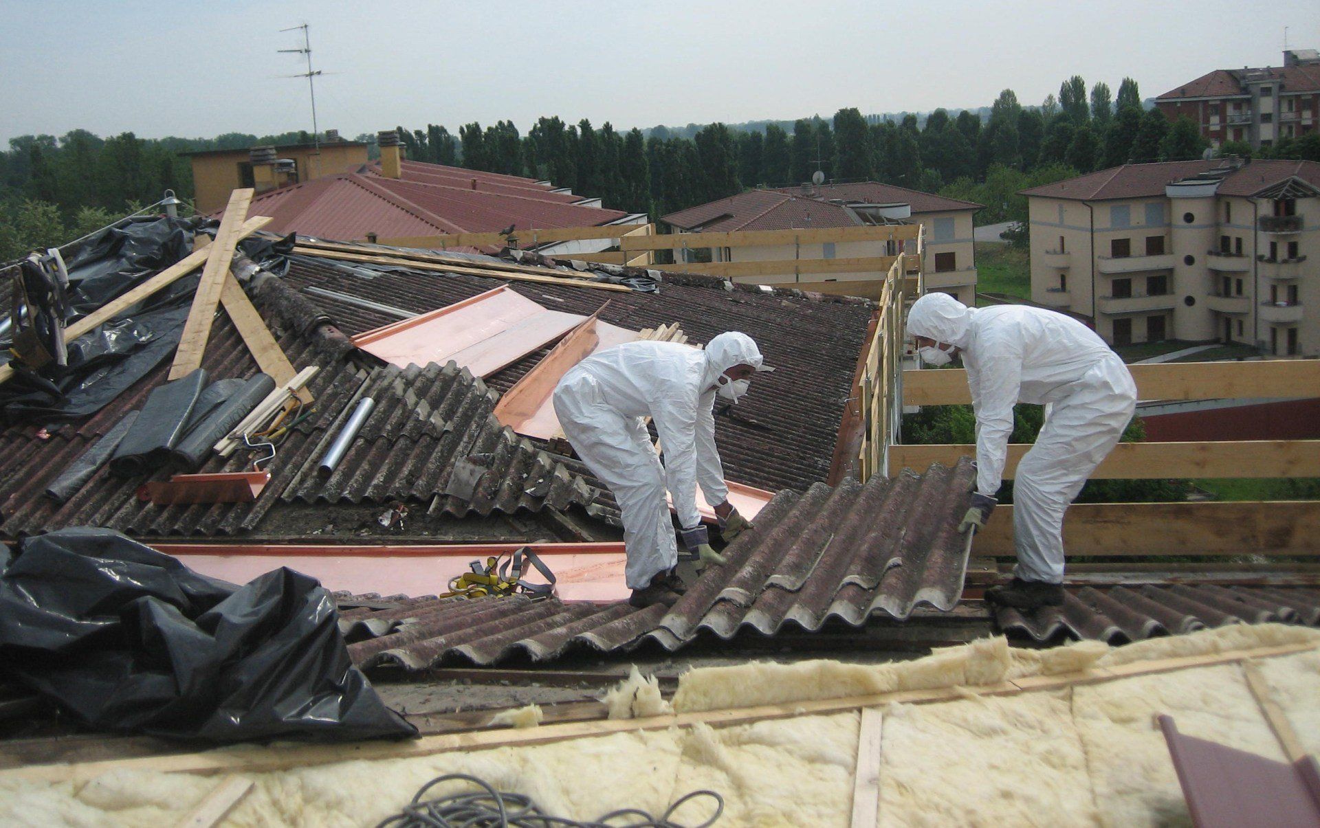 Due operai, con indosso tute e maschere protettive bianche, rimuovono con cura i pannelli ondulati del tetto di un edificio.
