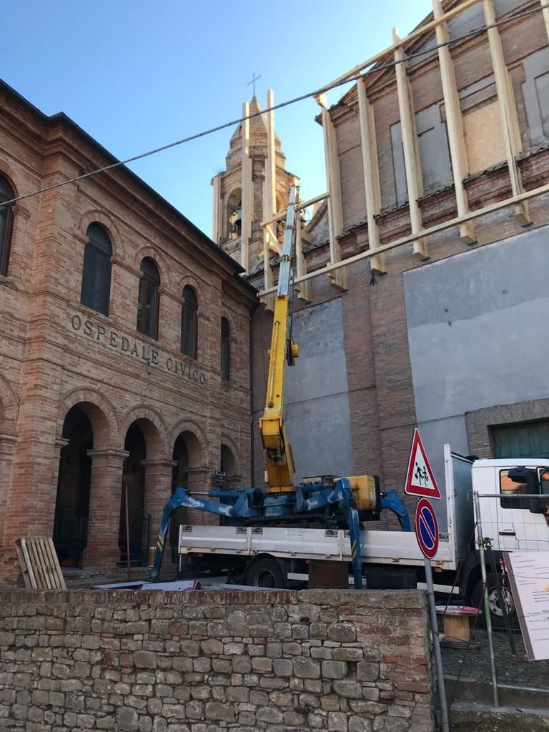 Una piattaforma aerea gialla montata su un camion è parcheggiata di fronte a un edificio storico in mattoni con campanile e impalcatura in legno.