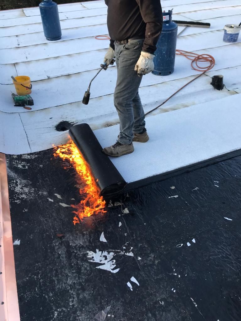 Un operaio utilizza una torcia a propano per fondere e incollare una membrana di copertura nera in rotoli su una superficie bianca.