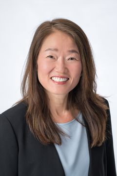 Woman with long brown hair, smiling, wearing a blue shirt and black blazer.