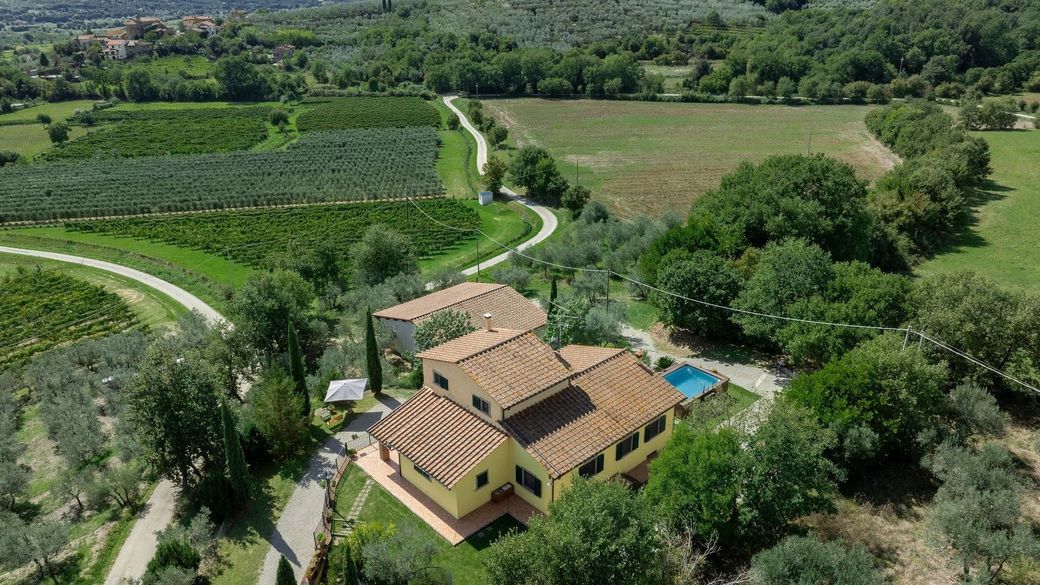 Yellow house with orange tile roof, set amidst green fields and trees, possibly rural Italy.