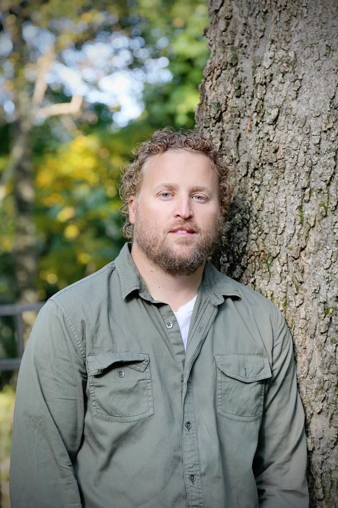 A woman in a green shirt and blue jeans is standing in front of a tree.