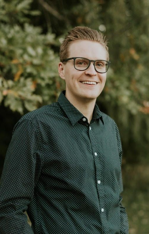 Man with glasses smiling, wearing a dark patterned shirt, standing outdoors with green foliage in the background.