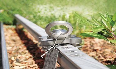 Anclaje de seguridad metálico con ojal en un riel metálico, para exteriores. Una etiqueta cuelga del ancla.