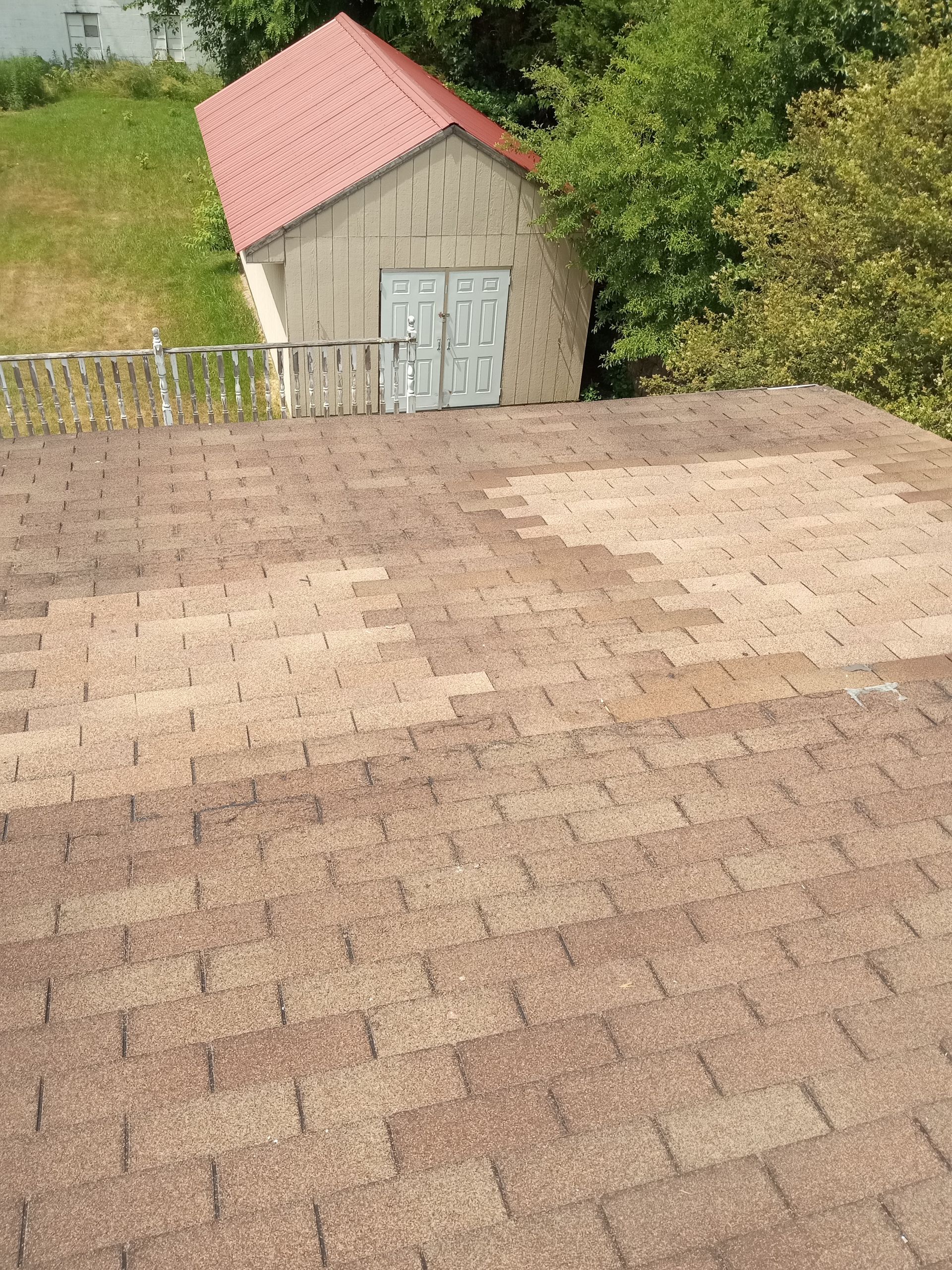 Brown shingle roof with faded patches, shed with red roof and green trees in background.