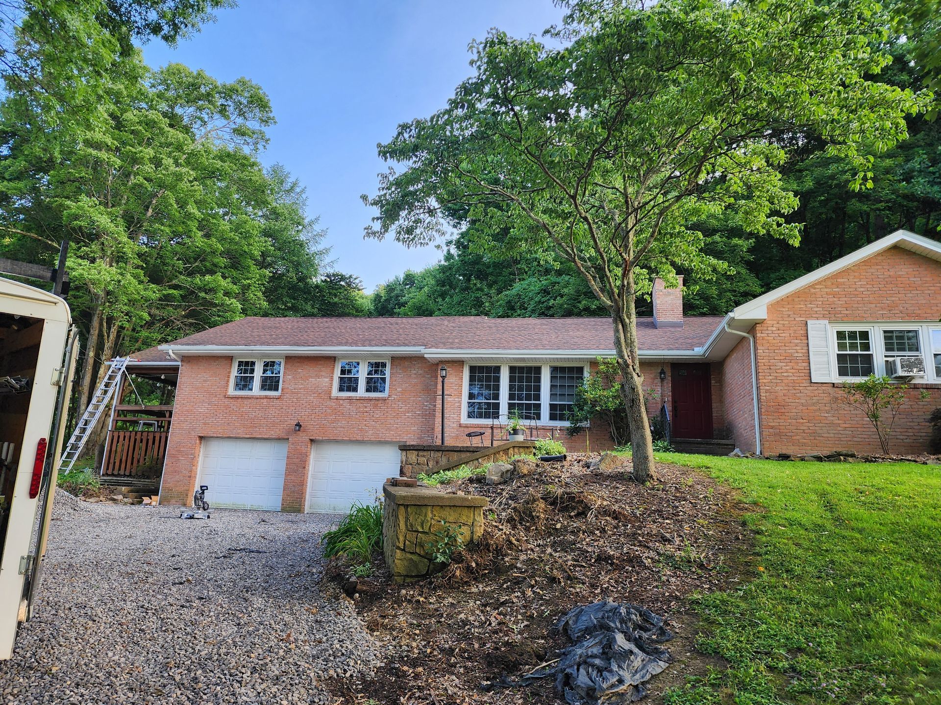 Brick house with two-car garage, red door, and gravel driveway. Surrounded by trees and greenery.