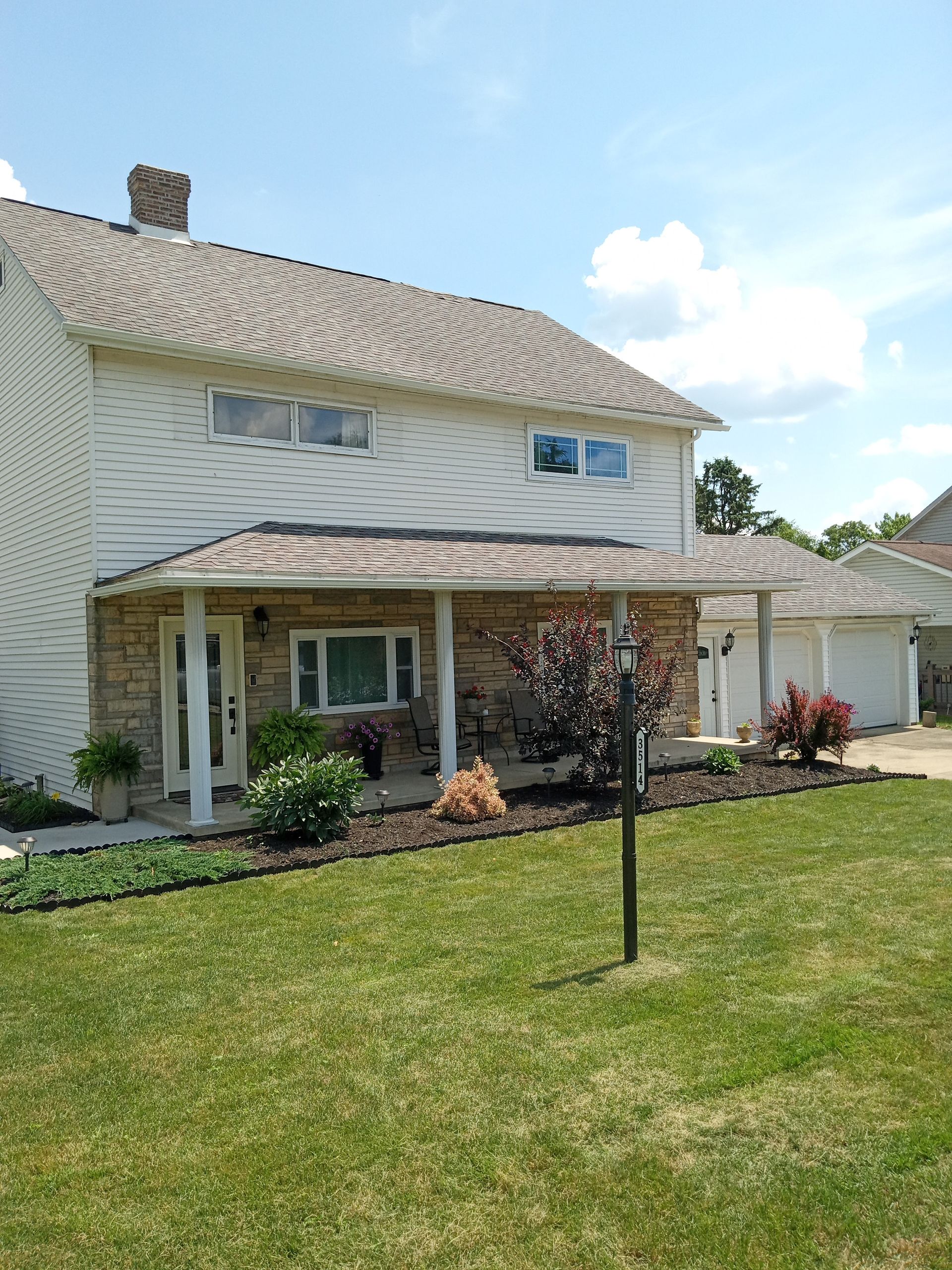 Two-story house with tan siding, stone facade, and a covered porch. A grassy lawn and a garage are visible.