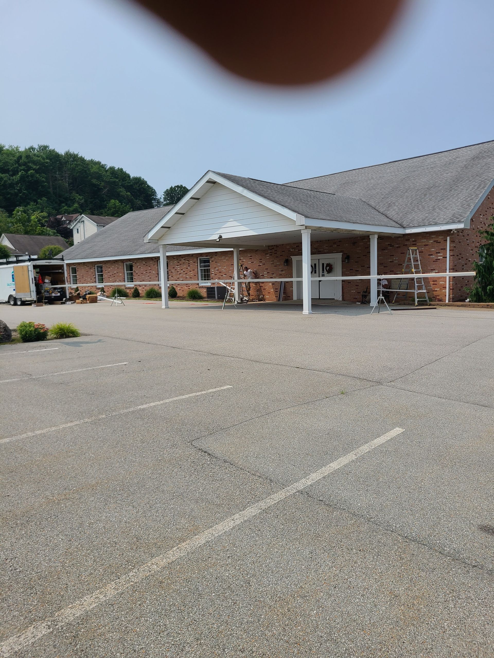 Building with brick facade and covered entrance, gravel parking lot, trees in background.