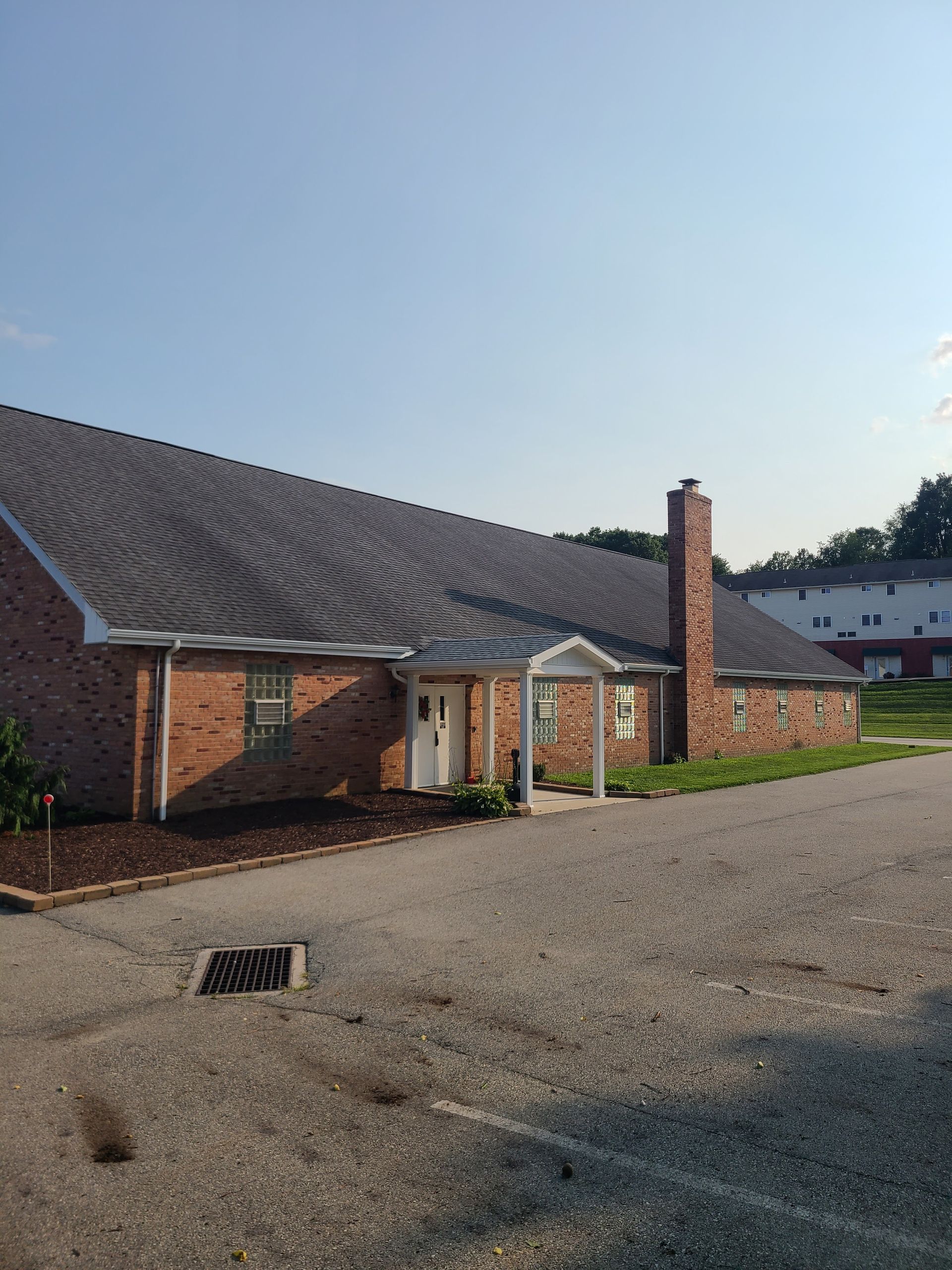 Brick building with dark roof, chimney, and entrance under an awning; asphalt driveway in foreground.