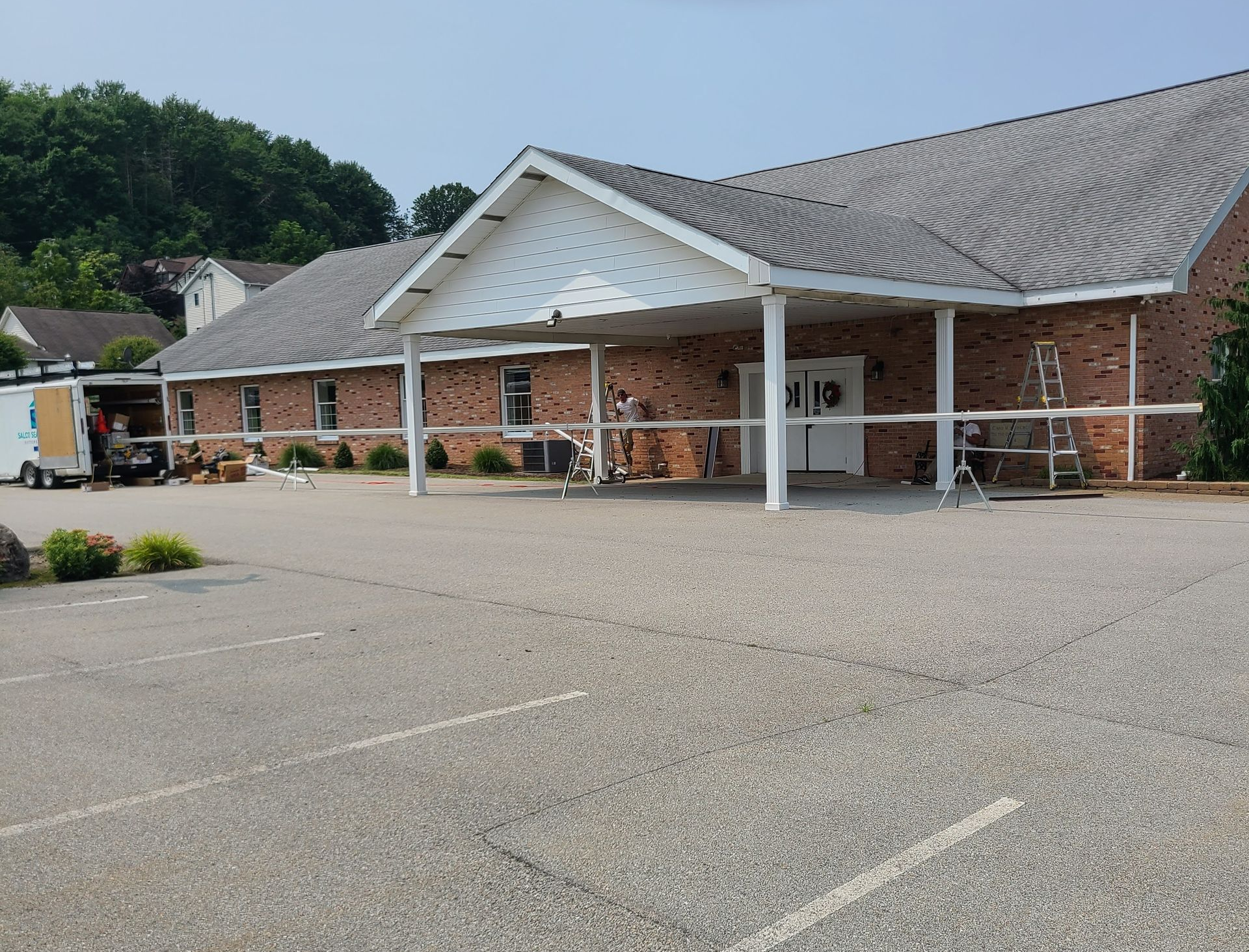 Exterior of a brick building with a covered entrance and a parking lot in front; work in progress.
