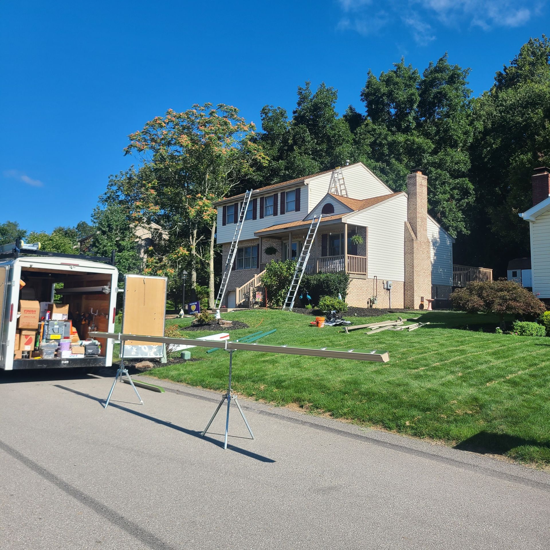 A two-story house with ladders and construction materials. A work van is parked in the driveway. Green lawn, blue sky.