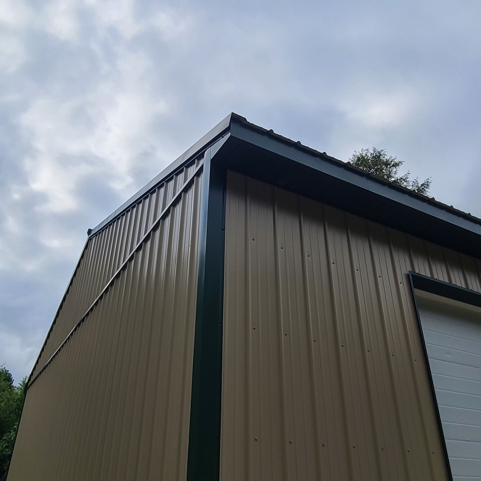 Tan metal building with dark green trim and a white garage door, against a cloudy sky.