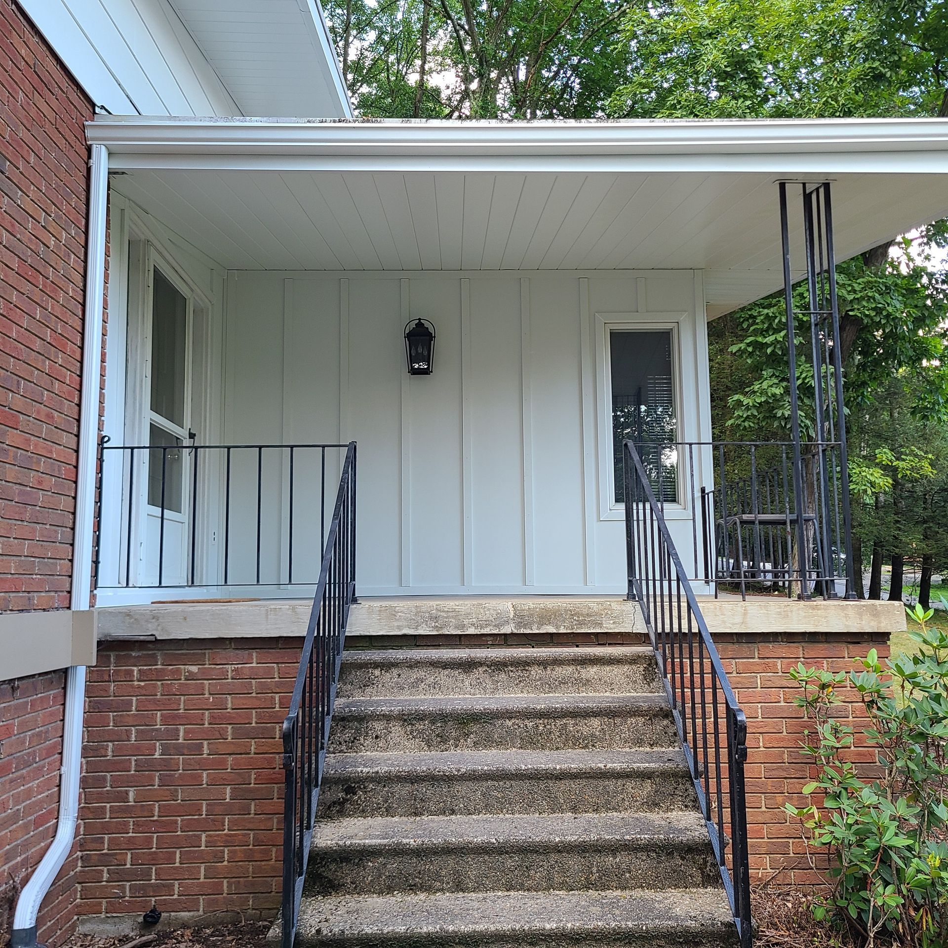 Brick house porch with steps and black railing. White siding and a black light fixture.