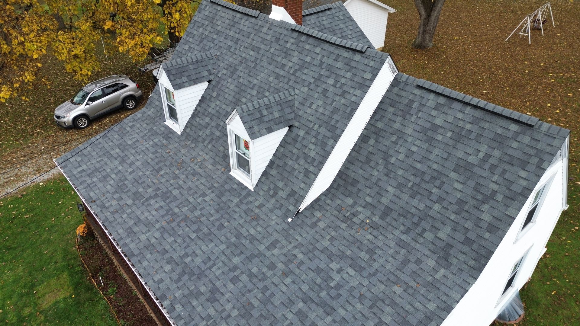 Overhead view of a house with a gray shingle roof, three white dormers, and a car in the yard.