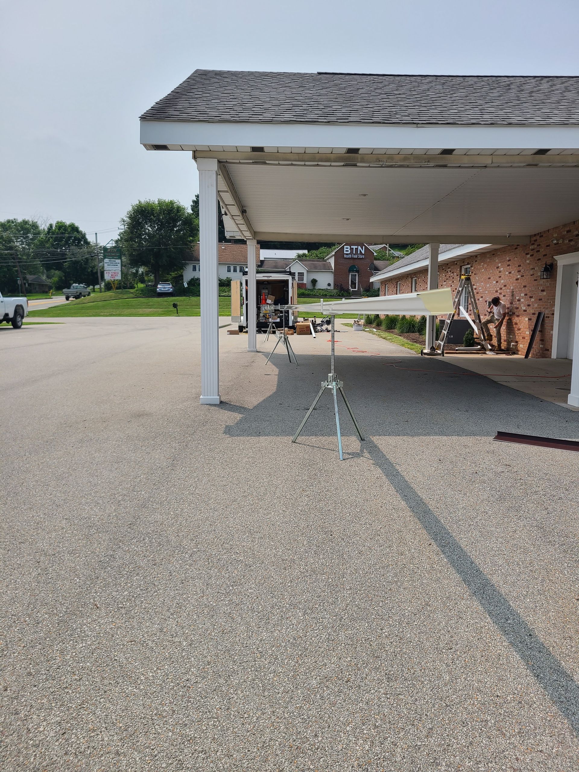 Carport with gravel parking lot. A few objects and a parked trailer are visible. Sunny day.