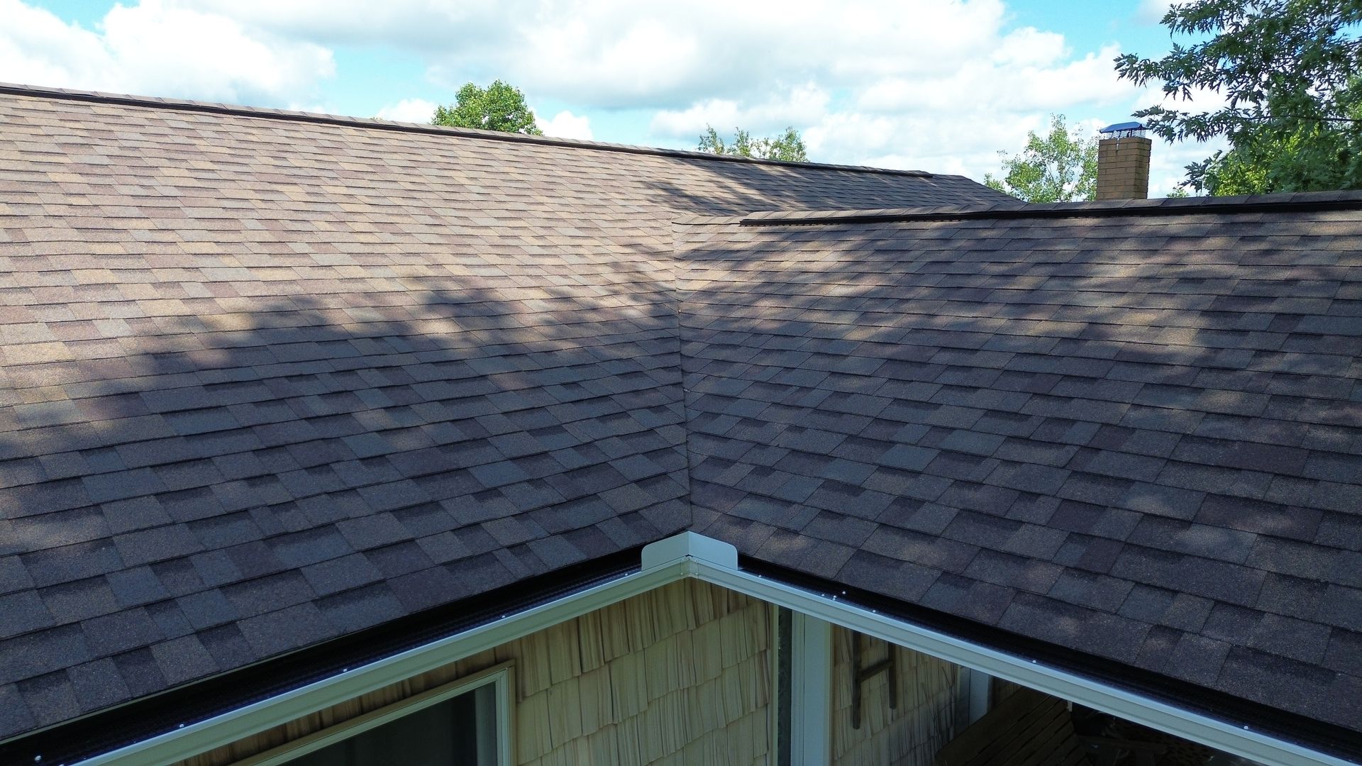 Brown shingle roof with a corner, shadows, and a glimpse of a house and trees.