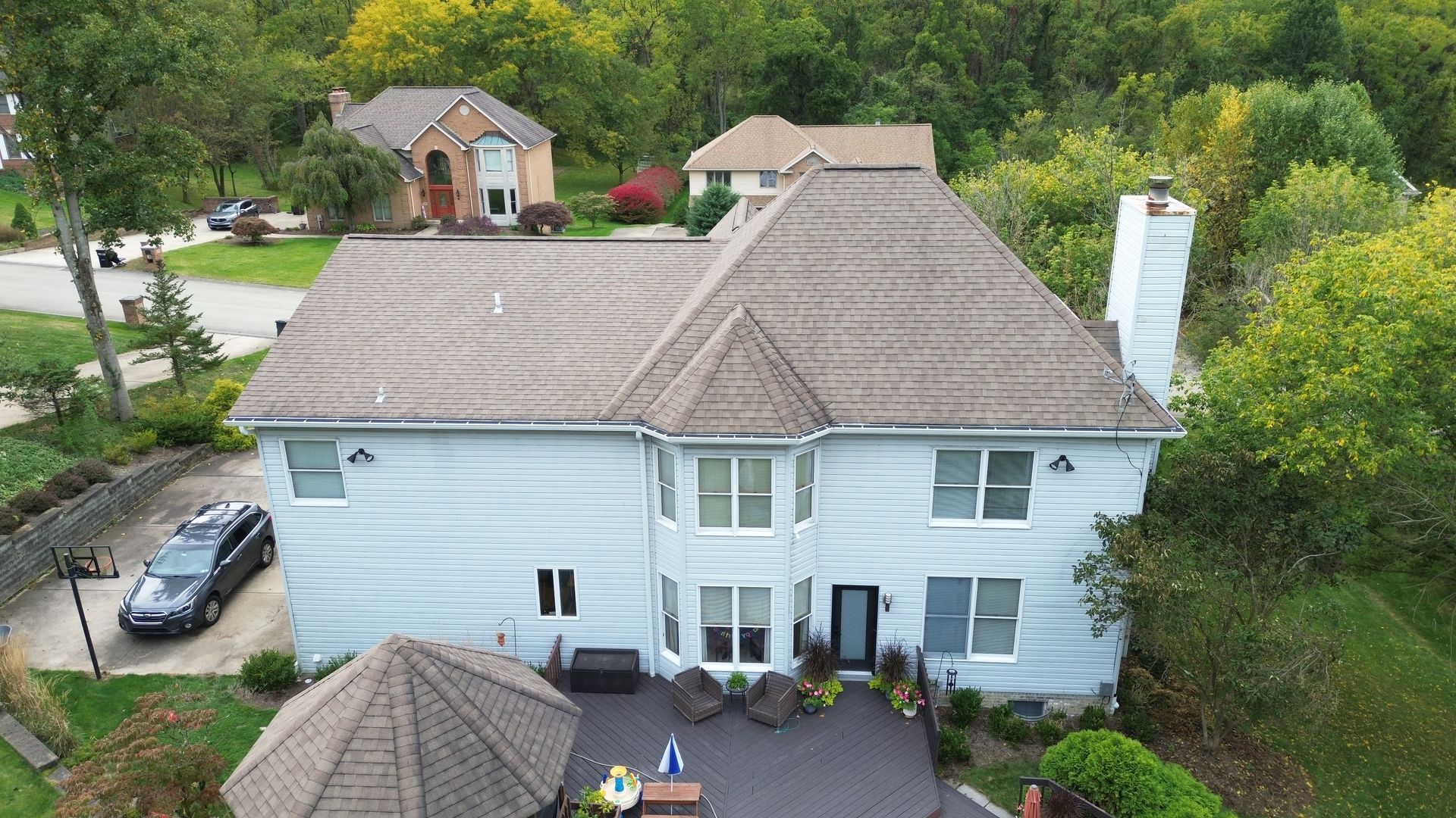 Aerial view of a light blue house with a brown roof and a black patio. A car is parked in the driveway.