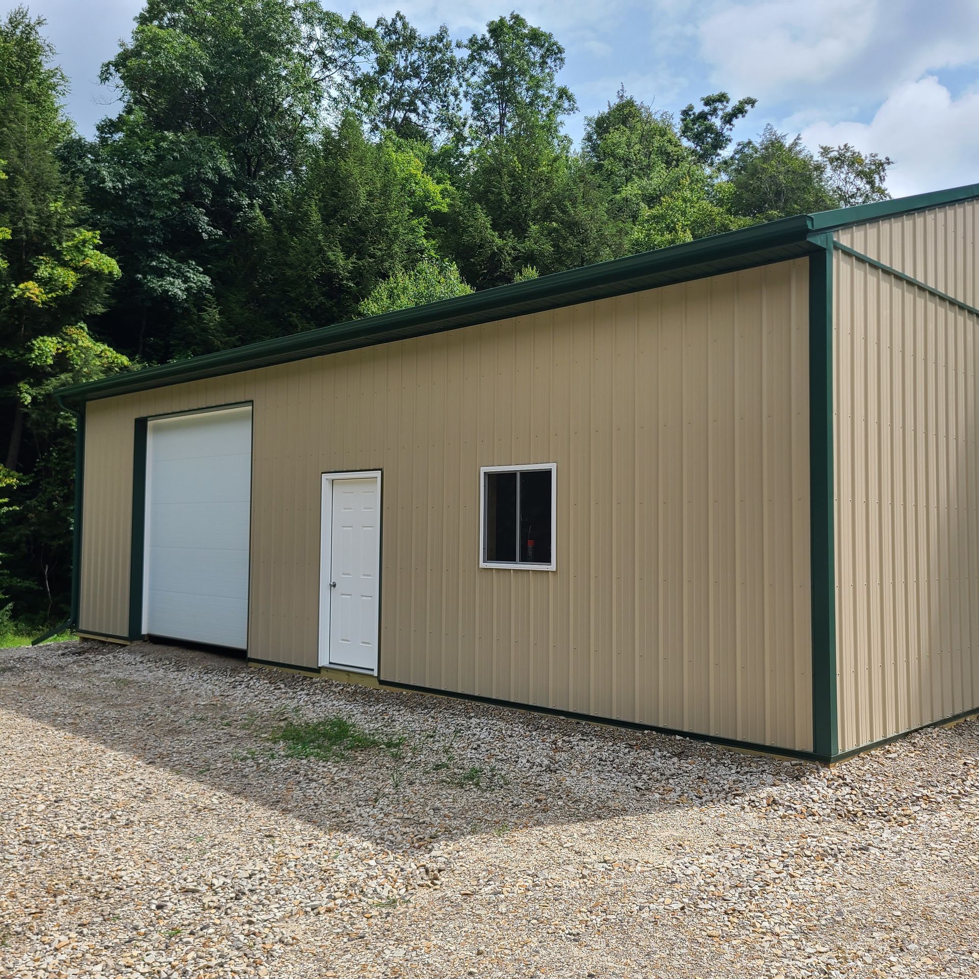 Tan metal building with green trim, roll-up door, small window, and white door; gravel driveway.