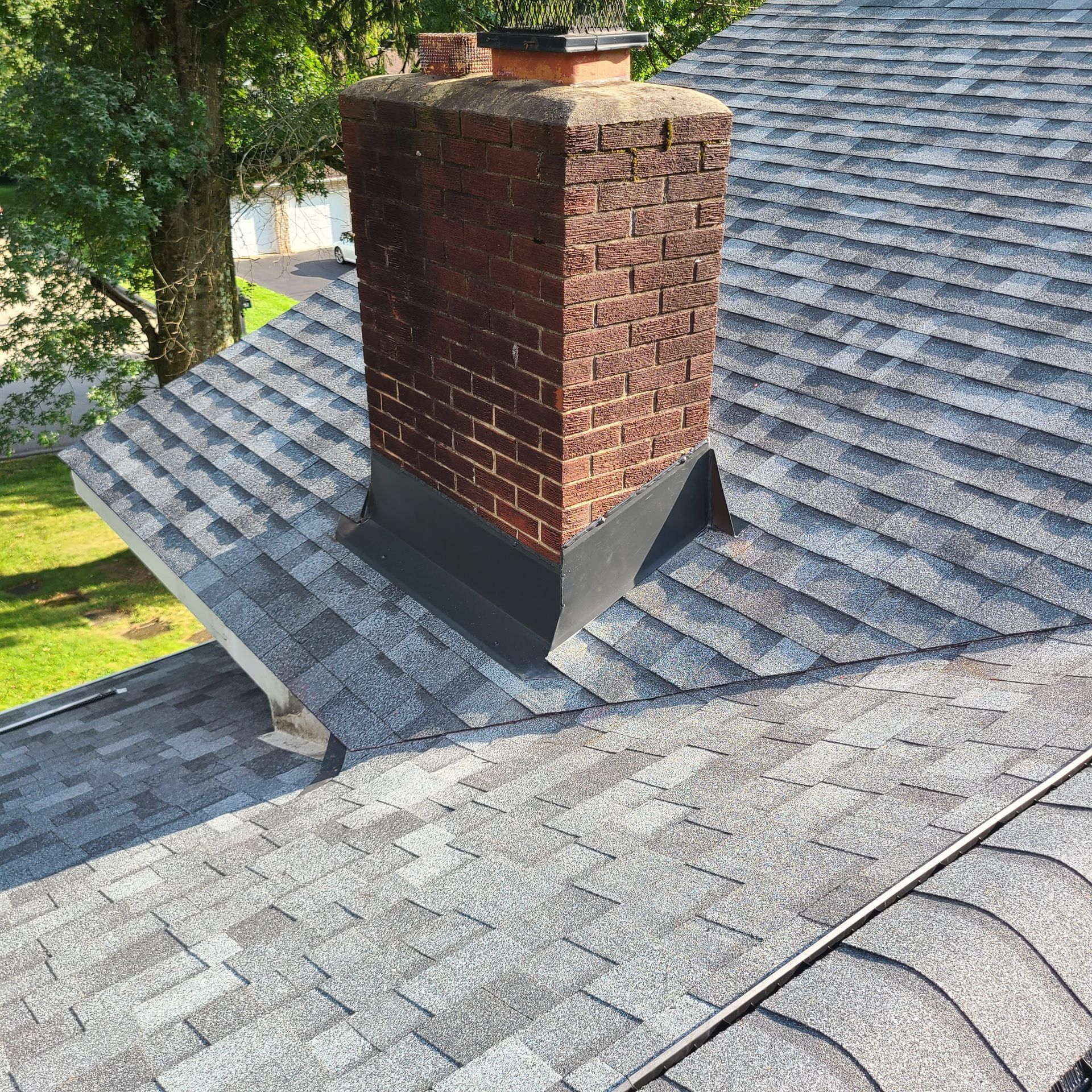 Brick chimney on a gray shingle roof, surrounded by green grass and trees.